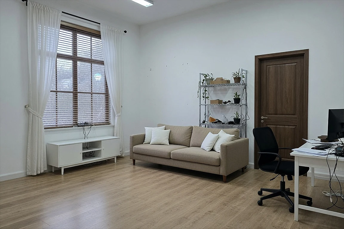 Basic creative studio before workspace interior transformation, with simple sofa, desk, visible cables, metal shelf, white walls, wood floor and underdesigned room potential.