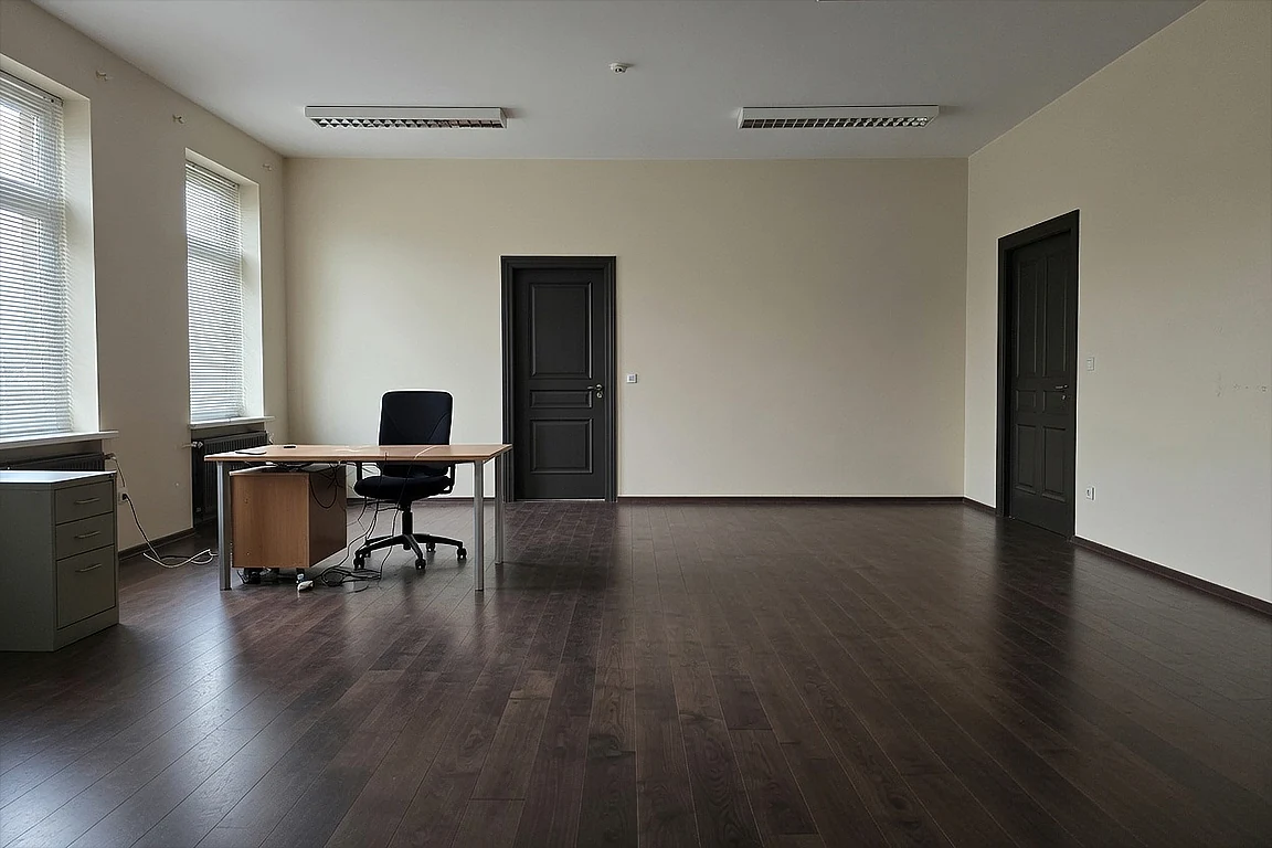 Large underdesigned room before red lounge transformation, with plain beige walls, dark wood floor, simple desk, office chair, fluorescent ceiling lights and unused dramatic potential.