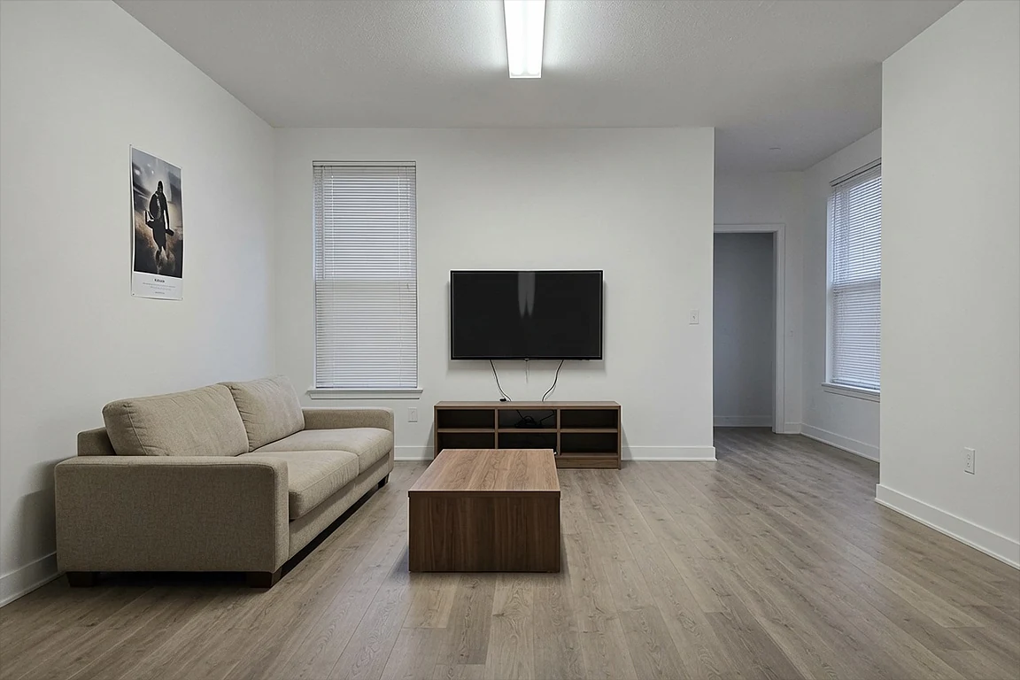 Underdesigned living room before AI interior design concept, with beige sofa, wall-mounted TV, simple coffee table, blinds, plain white walls, visible cables and cold ceiling light.