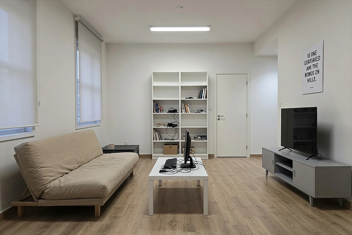 Basic living studio before AI room mood transformation, with simple sofa, TV stand, white shelves, coffee table, plain walls, cold ceiling light and generic apartment styling.