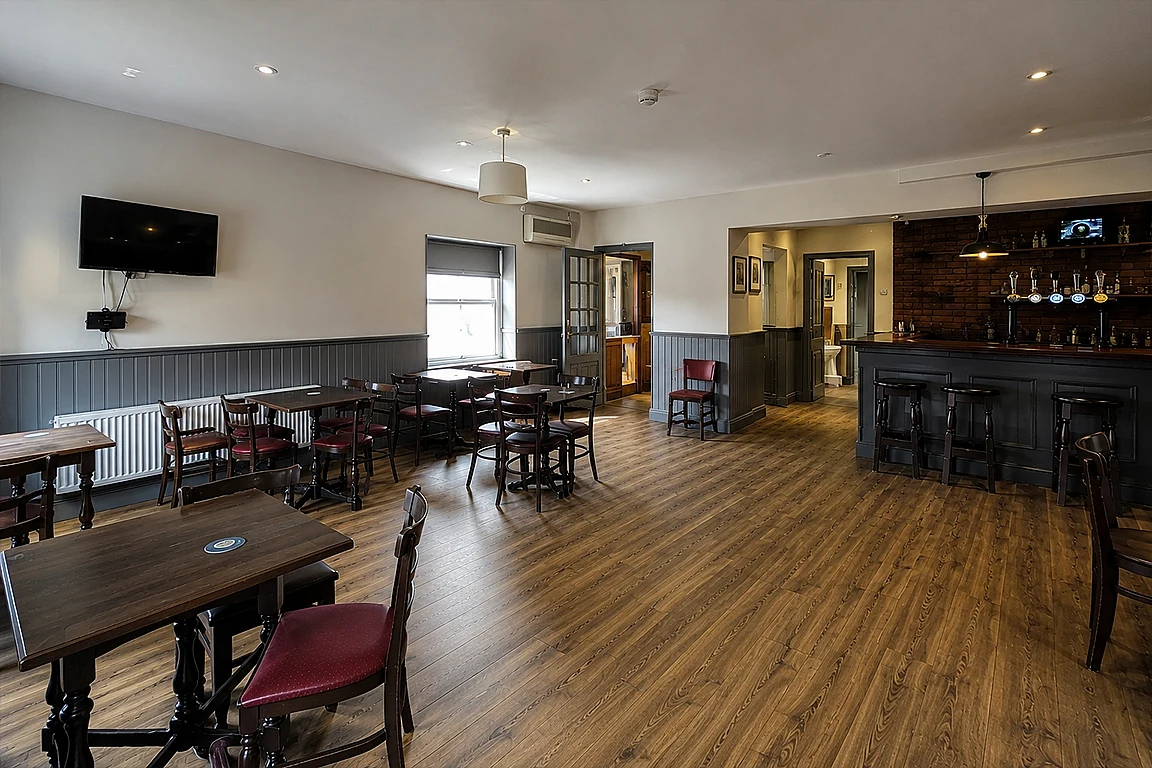 Small pub lounge before interior mood transformation, with wooden tables, bar counter, wall-mounted TV, plain ceiling lights, empty floor space and basic hospitality presentation.